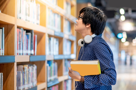 Young Asian man student choosing book in public library. Male researcher looking for textbook on bookshelves. Education research and self learning in university life conceptsの写真素材