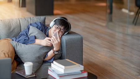 Young Asian man using smartphone for listening to music on mobile application, lying and relaxing on sofa during free time. Urban lifestyle in living space conceptの写真素材