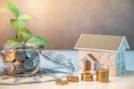 Real estate investment or property ladder. Home mortgage loan rate. Saving money for future concept. Plant growing out of coins in glass jar with dollar banknotes and house model on the tableの写真素材