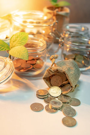 Coins spilling out of money bag with currency glass jars and green plant on the table. Saving money...