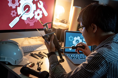 Asian technical engineer using screwdriver for repairing drone with computer and other tools on desk. Male technician fixing or maintenance drone. Unmanned aerial vehicle (UAV) photography conceptの写真素材