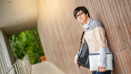 Young Asian man dressed in casual style carrying backpack and holding file and laptop computer in university. Happy male adult student in college.の写真素材