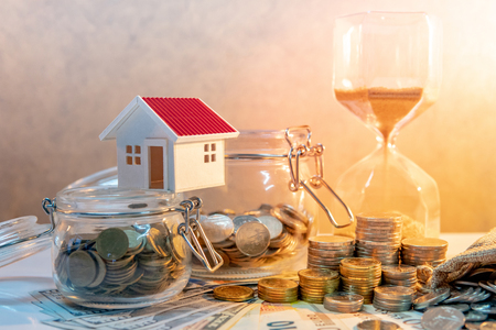 Saving money for retirement concept. Real estate or property investment. Home mortgage loan rate. Coin stack, money bag and currency glass jars on banknotes with hourglasses, house model on the table.の写真素材