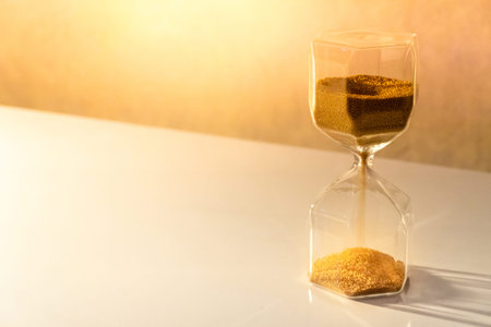 Gold sand running through the shape of modern hourglass on white table.Time passing and running out...