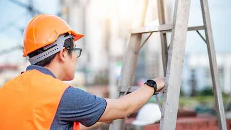 Young Asian maintenance worker man with orange safety helmet and vest carrying aluminium step ladder at construction site. Civil engineering, Architecture builder and building service conceptsの写真素材