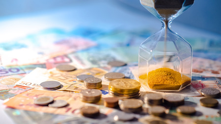 Sand running through the shape of hourglass on table with banknotes and coins of international currency. Time investment and retirement saving. Urgency countdown timer for business deadline conceptの写真素材