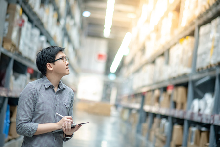 Young Asian man worker doing stocktaking of product in cardboard box on shelves in warehouse by using digital tablet and pen. Physical inventory count conceptの写真素材