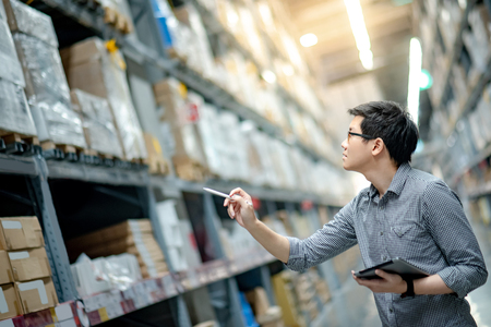 Young Asian man worker doing stocktaking of product in cardboard box on shelves in warehouse by using digital tablet and pen. Physical inventory count conceptの写真素材