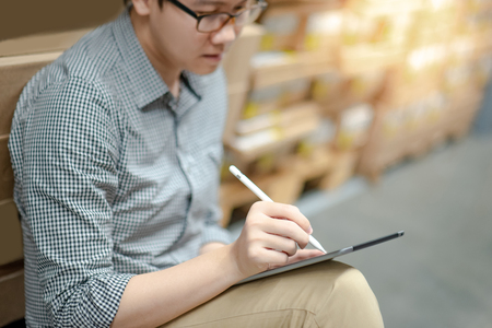 Young Asian man worker doing stocktaking of product in cardboard box on shelves in warehouse by using digital tablet and pen. Physical inventory count conceptの写真素材