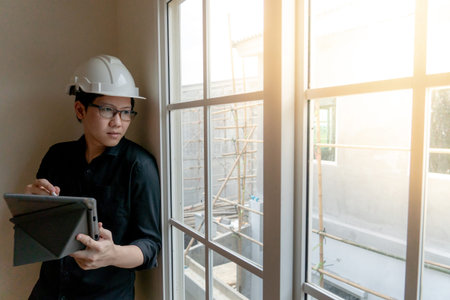 Young Asian engineer with helmet and eyeglasses using digital tablet looking through the window at...