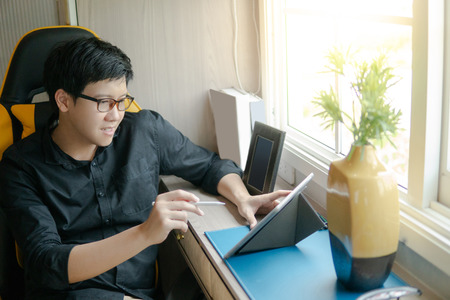 Young Asian business man with eyeglasses using digital tablet while sitting on the chair in working area. Home living lifestlyle with modern electronic gadget conceptの写真素材