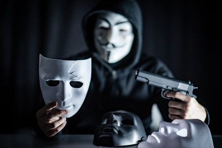 BANGKOK, THAILAND - SEPTEMBER 21, 2018 : Hoodie killer man wearing mystery mask holding gun preparing to shoot to white mask in his hand. Anonymous thief or burglar committing the crime.のeditorial素材