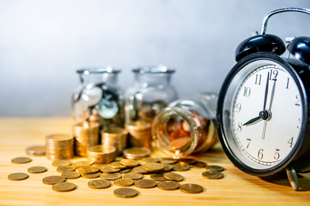 Clock with coins in currency glass jars and spilling on wooden table. Saving money for future retirement. Financial business growth. Time investment conceptの写真素材