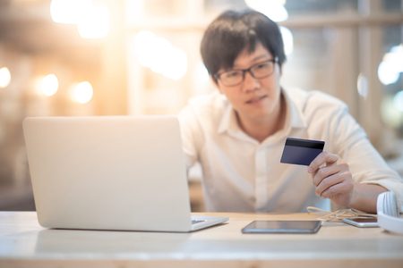 Young Asian man holding credit card using laptop computer for online shopping. Digital payment on E-commerce website. Electronic banking or cashless society conceptsの写真素材