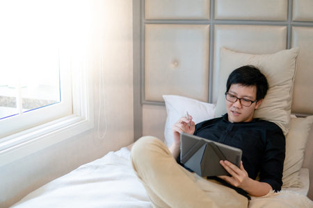 Young Asian business man using digital tablet and pen while lying on bed in cozy bedroom. Home living lifestyle with modern electronic gadget conceptの写真素材