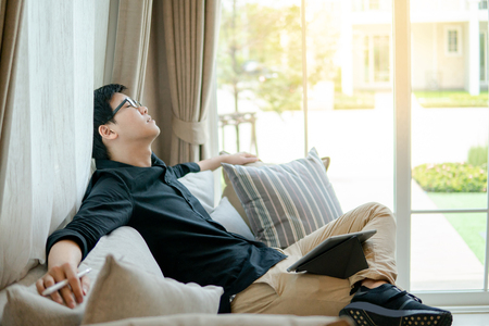 Young Asian business man sitting with digital tablet on sofa or couch in living room. Taking a rest at home. Cozy relaxing lifestyle conceptの写真素材
