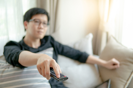 Close up of male Asian hand using remote controller while watching TV on sofa in home theater room. Home living and relaxing lifestyle concepts.の写真素材