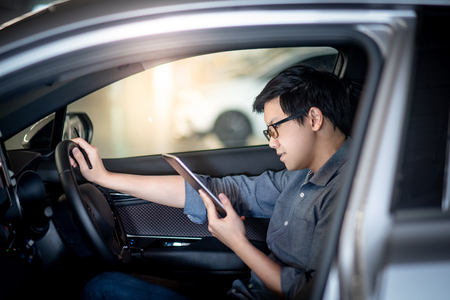 Young Asian businessman with glasses reading news on digital tablet while sitting on driver seat in his car. Business and technology conceptの写真素材