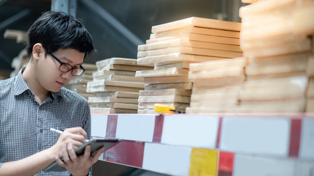 Young Asian man worker doing stocktaking of product in cardboard box on shelves in warehouse by using digital tablet and pen. Physical inventory count conceptの写真素材