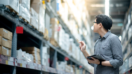 Young Asian man worker doing stocktaking of product in cardboard box on shelves in warehouse by using digital tablet and pen. Physical inventory count conceptの写真素材
