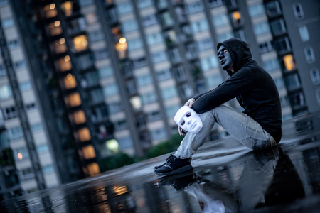 Reflection of mystery hoodie man with black mask holding white mask sitting in the rain on rooftop of abandoned building. Bipolar disorder or Major depressive disorder. Depression conceptの写真素材