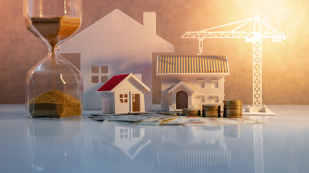 Real estate or property development. Construction business investment concept. Home mortgage loan rate. Coin stack on international banknotes with hourglasses, house and crane models on the table.の写真素材