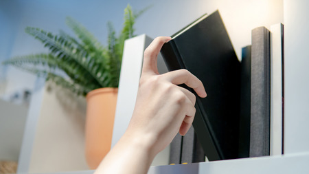 Male hand choosing and picking black book from bookshelf in public library. Education research and self learning in university life conceptsの写真素材