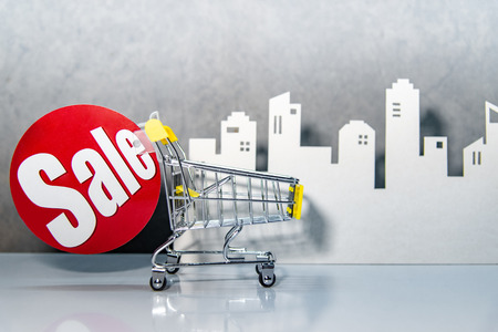 Mini shopping cart or shopping trolley with red sale label and city background on the table. Supermarket grocery push cart. Property or real estate on sale.の写真素材