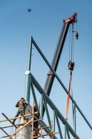 Male construction workers standing on wooden scaffolding welding steel truss structure on concrete column at construction site. Wide span building construction process with drone taking photo overhead.の写真素材
