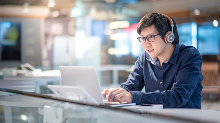Asian business man listening to music by headphones while working with laptop computer in coworking space. Freelance or digital nomad lifestyle. Online shopping or E-commerce social media on internetの写真素材