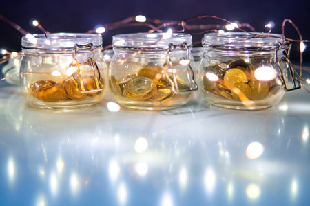 Gold coins in currency glass jars with decorative Christmas lights on glossy table. Saving money for...