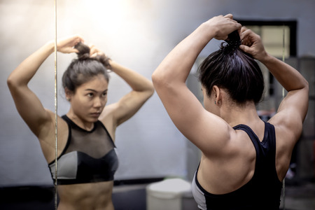 Asian athlete girl in sportswear looking into the mirror while tying her hair in fitness gym. Strong and confidence conceptsの写真素材