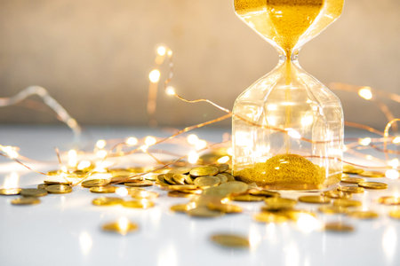 Gold sand running through the shape of hourglass with coins and decorative light on the table....