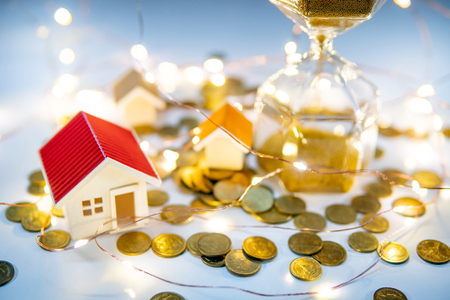 Real estate investment concept. Property marketing during festive holiday season. Hourglass with gold coins, house models and decorative lights on the table. Saving money for retirement.の写真素材
