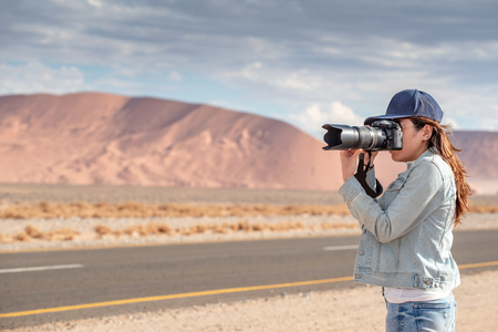 Young Asian woman traveler and photographer holding camera taking photo of sand dune near the road in Namib desert of Namibia, Africa. Travel photography conceptの写真素材