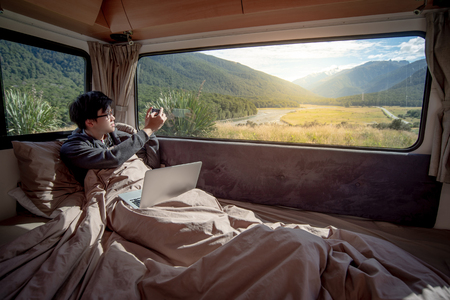 Asian man traveler enjoy taking photo of mountain scenery through the window by smartphone while staying in the blanket in camper van in the morning. Road trip in summer of South Island, New Zealand.の写真素材