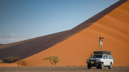 Young Asian man traveler and photographer standing on camper car near orange sand dune. Desert road trip travel conceptの写真素材
