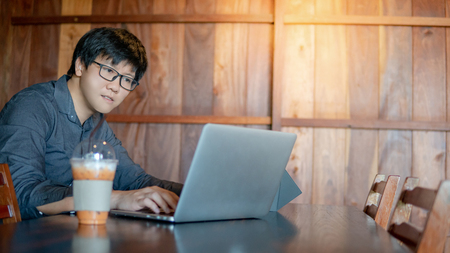 Male Asian businessman wearing glasses working with modern laptop computer on wooden table in the cafe. Work smart in digital age. Internet of things (IOT) conceptの写真素材