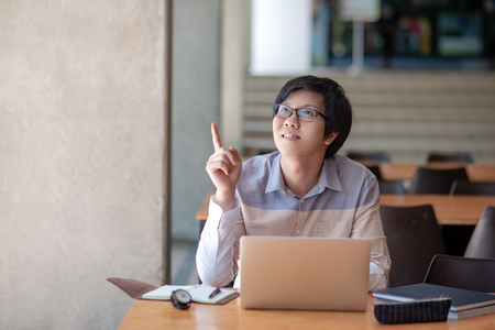 Young Asian man university student with glasses pointing finger up while using laptop computer in the college library. Education opportunity or scholarship. Idea and inspiration conceptsの写真素材