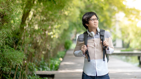 Confident Asian man university student with glasses and headphones smiling and holding backpack in the green park of college. Happy life in campus. Education opportunity and scholarship conceptの写真素材