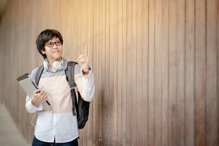 Asian man university student with glasses, haedphones and backpack pointing finger up and holding laptop computer and file in campus building corridor. Education opportunity or scholarship conceptsの写真素材