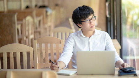 Young Asian business man with glasses working with laptop computer on wooden table looking outside...