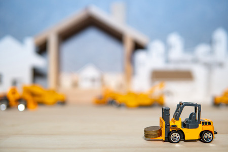Miniature yellow forklift model lifting gold coins stacked on wooden table with blurred house frame and city in the background. Warehouse logistics business or construction industry conceptの写真素材