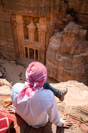 Asian man traveler sitting on carpet viewpoint in Petra ancient city looking at the Treasury or Al-khazneh, famous travel destination of Jordan and one of seven wonders.の写真素材