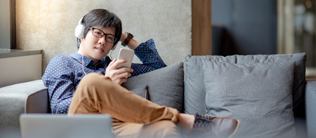 Young Asian man using smartphone for listening to music on mobile application, sitting and relaxing on sofa during free time. Urban lifestyle in living space conceptの写真素材