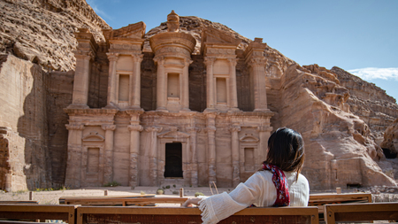 Asian woman tourist in white dress sitting and looking at Ad Deir or El Deir, the monument carved out of rock in the ancient city of Petra, Jordan.の写真素材
