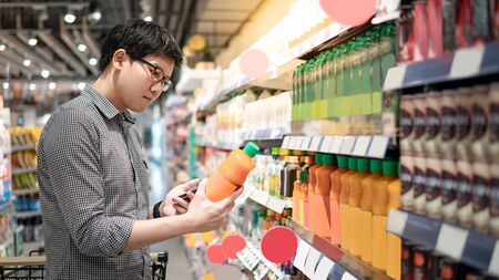 Asian man choosing orange juice in supermarket using smartphone to check shopping list. Male shopper with shopping cart selecting beverage bottle product in grocery store.の写真素材