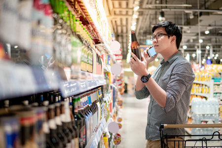 Asian man using smartphone while shopping beer in supermarket. Male shopper with shopping cart...