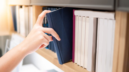 Male hand choosing and picking blue book from wooden bookshelf in public library. Education research and self learning in university life conceptsの写真素材