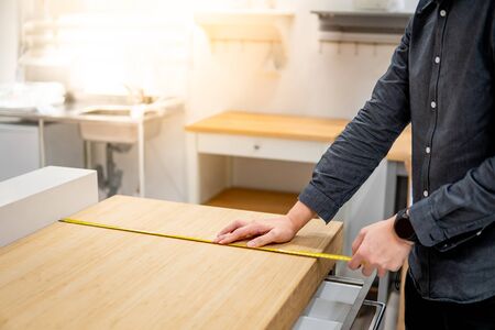 Asian man interior designer using tape measure for measuring size of wooden countertop in modern kitchen showroom in furniture store. Shopping material design for home improvement.の写真素材